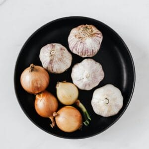 Overhead of raw unpeeled aromatic garlic and yellow onions with one regrowing onion bulb on black ceramic plate placed on white marble background