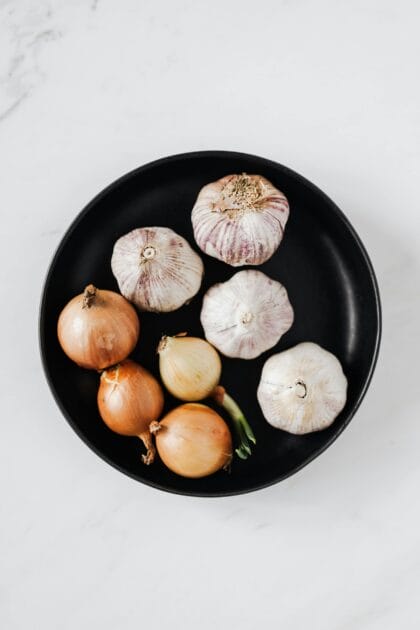 Overhead of raw unpeeled aromatic garlic and yellow onions with one regrowing onion bulb on black ceramic plate placed on white marble background