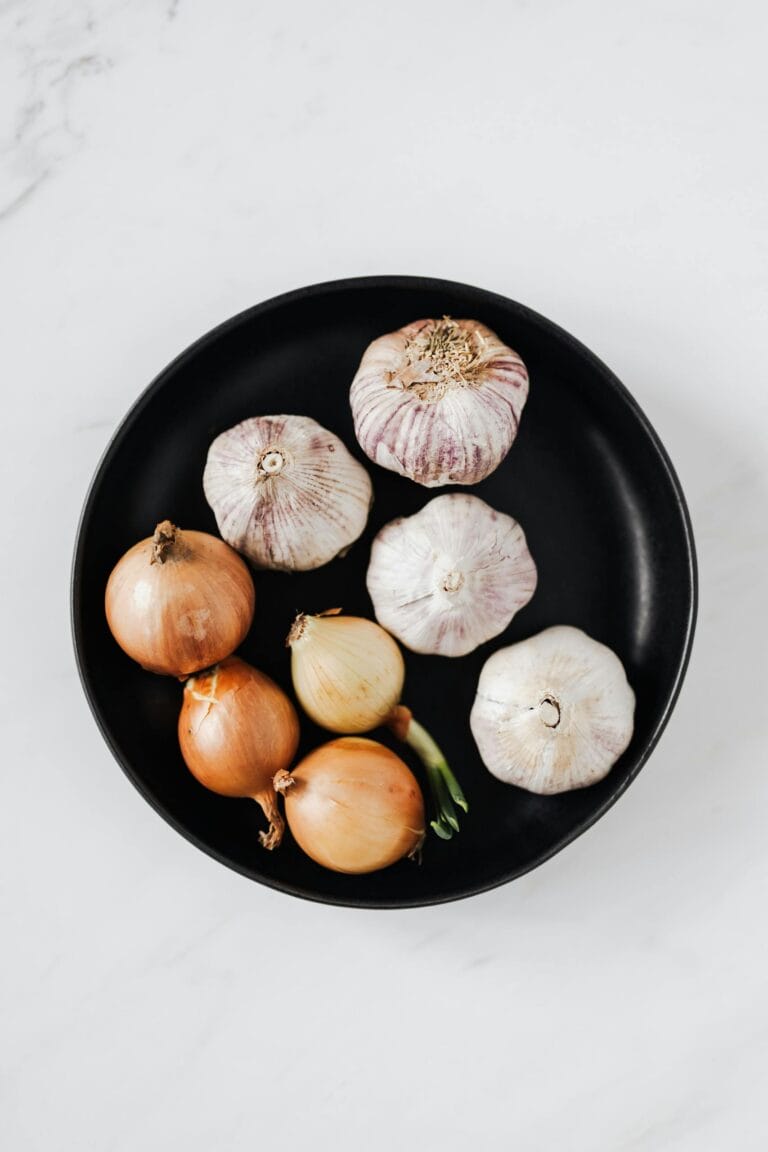 Overhead of raw unpeeled aromatic garlic and yellow onions with one regrowing onion bulb on black ceramic plate placed on white marble background